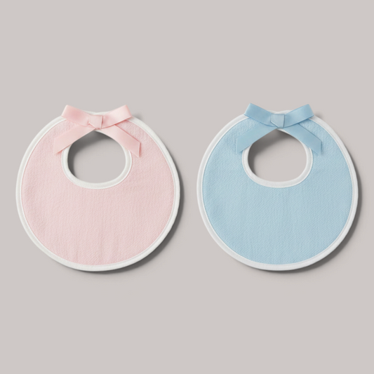 Dos baberos NAVIA de algodón suave, uno rosa y otro azul, con ribete blanco y un delicado lazo decorativo.